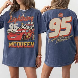 May include: A blue t-shirt featuring a graphic of Lightning McQueen from the Cars movie. The front design includes the text "Lightning 95 McQueen" with a red race car. The back of the shirt displays a large "95" and "Lightning McQueen".