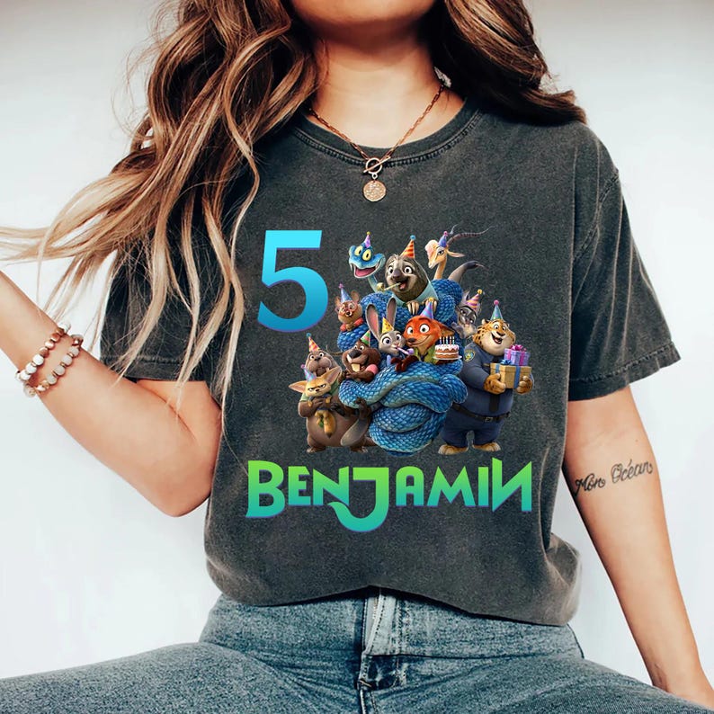 May include: Dark gray t-shirt featuring a colorful graphic of cartoon characters, the number "5", and the name "Benjamin" in green. The design includes various animal characters with party hats and a birthday cake. The shirt is a casual style.