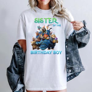 May include: White t-shirt with a colorful graphic of cartoon animals and the text "SISTER OF THE BIRTHDAY BOY" in green and blue. The design features various animal characters in party hats.