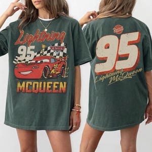 May include: Dark green t-shirt featuring a graphic of Lightning McQueen from the Cars movie. The front design includes the text "Lightning 95 McQueen" with a red race car. The back displays "95 Lightning McQueen".