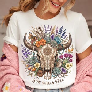 May include: White t-shirt featuring a detailed illustration of a cow skull adorned with flowers, feathers, and a butterfly. The design includes the text "Stay Wild & Free" in a decorative font. The shirt has short sleeves.