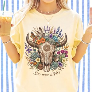 May include: A light yellow t-shirt featuring a detailed illustration of a cow skull adorned with flowers, feathers, and a butterfly. The design includes the text "Stay Wild & Free" in a decorative font. The shirt is a relaxed fit.