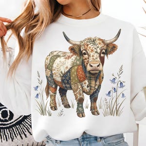 Könnte beinhalten: Weißes Sweatshirt mit einer Aquarellillustration einer Highland-Kuh. Die Kuh hat ein Patchwork-Design mit floralen und karierten Mustern in Blau, Grün und Braun. Glockenblumen und hohes Gras umgeben die Kuh.