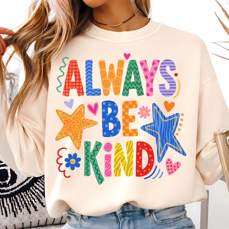May include: Cream-colored sweatshirt with the words "ALWAYS BE KIND" in colorful, whimsical lettering. The design includes stars, hearts, and floral accents. A casual and cheerful top.