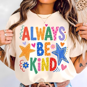 以下が含まれることがあります： クリーム色のTシャツ。カラフルで模様入りの文字で「ALWAYS BE KIND」と書かれ、星、ハート、花が飾られています。明るい色と遊び心のあるフォントが特徴で、陽気で前向きなメッセージを伝えています。