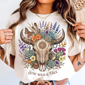 May include: Cream-colored t-shirt featuring a detailed illustration of a bull skull adorned with flowers, feathers, and a butterfly. The design includes the text "Stay Wild & Free" in a decorative font, perfect for a bohemian style.