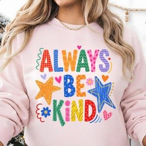May include: A light pink sweatshirt with the words "ALWAYS BE KIND" in colorful, whimsical lettering. The design includes stars, hearts, and floral accents, creating a cheerful and positive vibe. A casual and comfortable top.