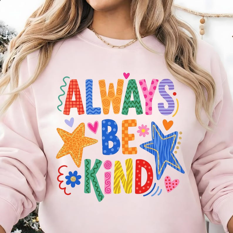 May include: A light pink sweatshirt with the words "ALWAYS BE KIND" in colorful, whimsical lettering. The design includes stars, hearts, and floral accents, creating a cheerful and positive vibe. A casual and comfortable top.