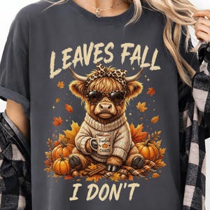 May include: A charcoal gray t-shirt featuring a graphic of a Highland cow wearing a leopard print headband and sunglasses, dressed in a sweater, holding a mug labeled "Fueled by Chaos," surrounded by pumpkins and leaves with the text "LEAVES FALL I DON'T."