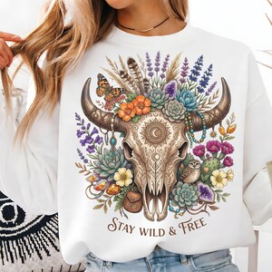May include: White sweatshirt featuring a detailed illustration of a bull skull adorned with flowers, feathers, and a butterfly. The design includes the text "Stay Wild & Free" in a decorative font. The artwork incorporates various colors.