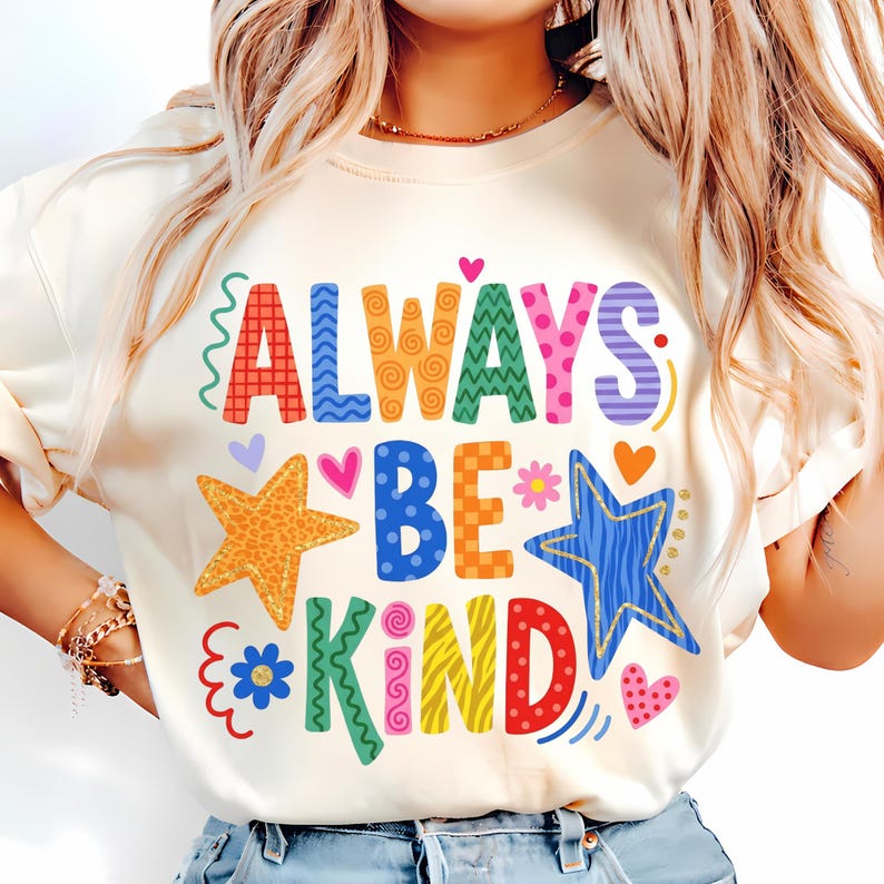 May include: Cream-colored t-shirt with the words "ALWAYS BE KIND" in colorful, whimsical lettering. The design includes stars, hearts, and flowers in various bright colors. The shirt has long sleeves.