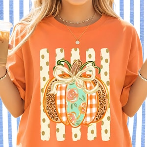 May include: An orange t-shirt featuring a central graphic of a pumpkin decorated with a white bow, orange and white gingham patterns, leopard print sections, and floral motifs, set against a background of vertical cream stripes with gold dots.