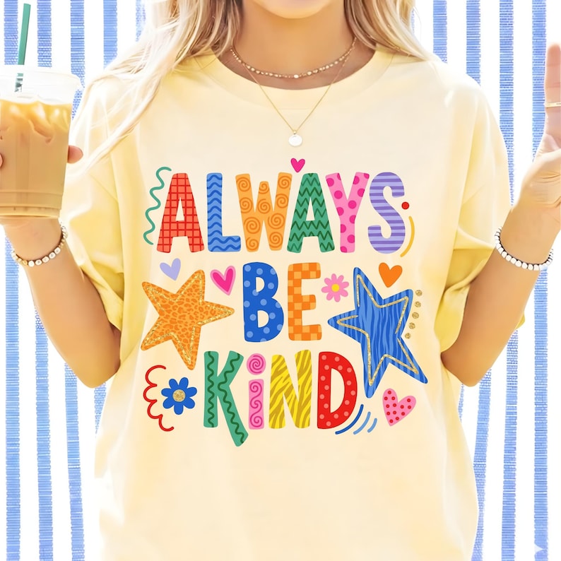 May include: A light yellow t-shirt with the words "ALWAYS BE KIND" in colorful, whimsical lettering. The design includes stars, hearts, and floral accents. The shirt has a relaxed fit.