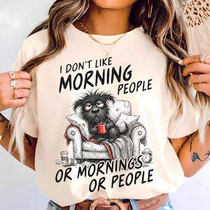 Könnte beinhalten: Cremefarbenes T-Shirt mit dem Text "I DON'T LIKE MORNING PEOPLE OR MORNINGS OR PEOPLE". Ein Cartoon-Hund sitzt in einem Stuhl und hält eine rote Tasse. Eine Kaffeetasse und ein Wecker sind ebenfalls zu sehen.
