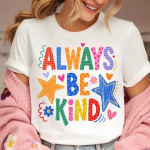 May include: White t-shirt with the words "ALWAYS BE KIND" in colorful, patterned letters. The design includes stars, hearts, and floral accents in various colors, creating a cheerful and positive vibe.