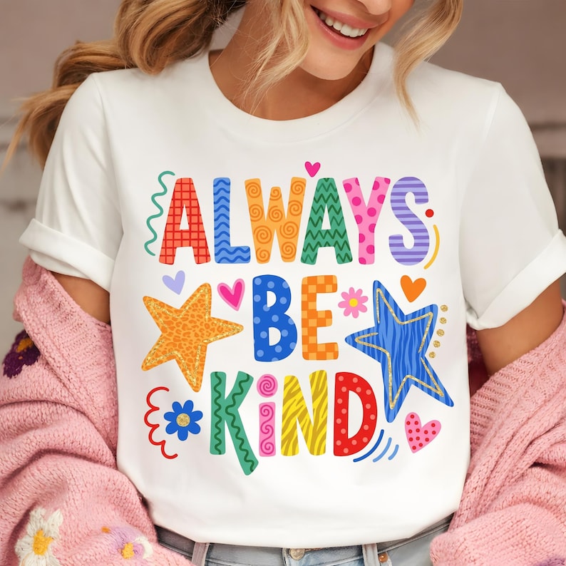 May include: White t-shirt with the words "ALWAYS BE KIND" in colorful, patterned letters. The design includes stars, hearts, and floral accents in various colors, creating a cheerful and positive vibe.