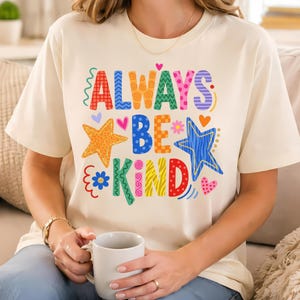 May include: Cream-colored t-shirt with the words "ALWAYS BE KIND" in colorful, playful lettering. The design includes stars, hearts, and flowers. The shirt is a casual, everyday wear item.