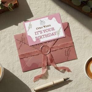 May include: A birthday-themed gift set featuring a mauve envelope with a ribbon and wax seal, a card that reads "Open When It's Your Birthday", a pen, and two cups of coffee with heart-shaped latte art.