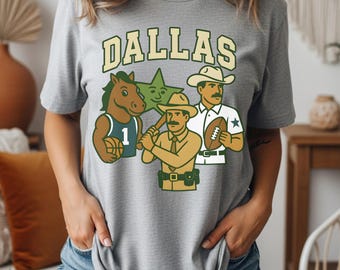 Puede incluir: Una camiseta gris con la palabra "DALLAS" en una fuente verde y beige. La camiseta presenta ilustraciones de dibujos animados de un caballo, una estrella, un jugador de béisbol y un jugador de fútbol, todo en un estilo vintage.