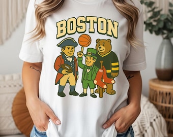 Puede incluir: Camiseta blanca con la palabra "BOSTON" sobre ilustraciones de dibujos animados de un patriota, un duende sosteniendo una pelota de baloncesto y un oso con una camiseta. El patriota sostiene un balón de fútbol y el oso una media.