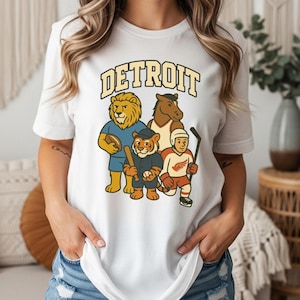 May include: White t-shirt featuring the word "DETROIT" in a vintage font above cartoon mascots of Detroit sports teams: a lion, a horse, a tiger, and a hockey player. The shirt is a classic crew neck.