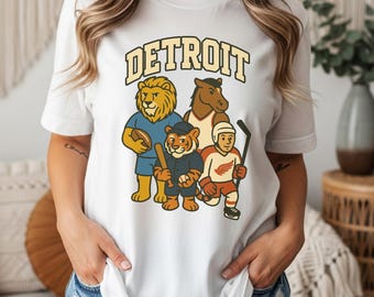 Puede incluir: Camiseta blanca con la palabra "DETROIT" en una fuente retro. Presenta mascotas de dibujos animados de equipos deportivos de Detroit: un león, un caballo, un tigre y un jugador de hockey. Cuello redondo.