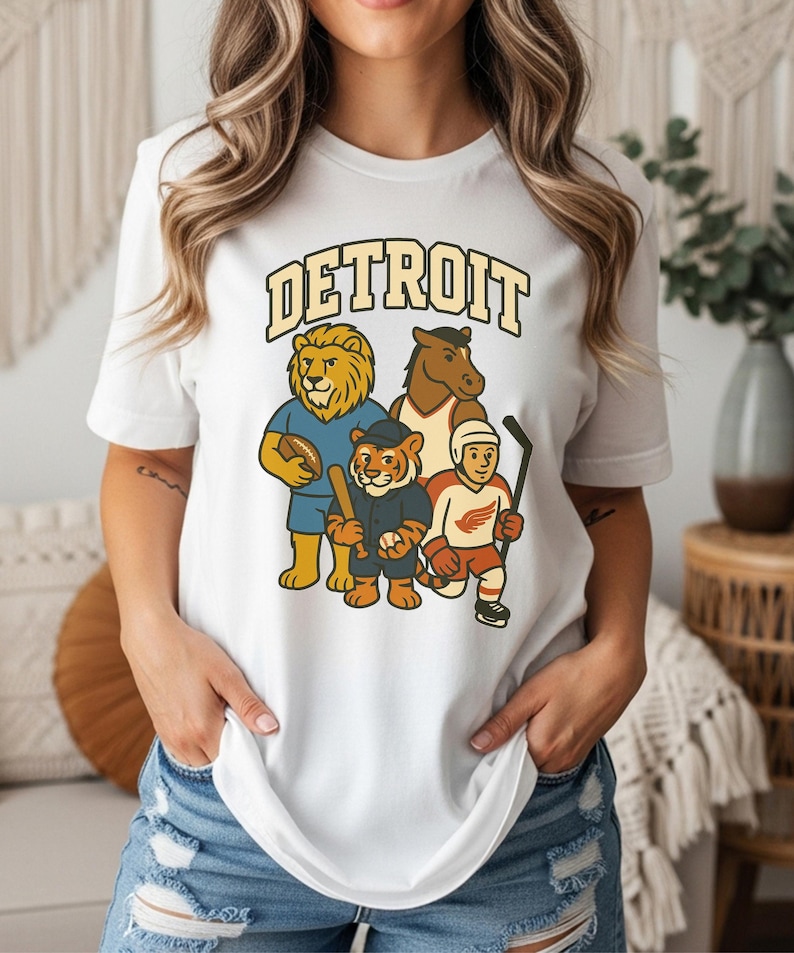 May include: White t-shirt featuring the word "DETROIT" in a vintage font above cartoon mascots of Detroit sports teams: a lion, a horse, a tiger, and a hockey player. The shirt is a classic crew neck.