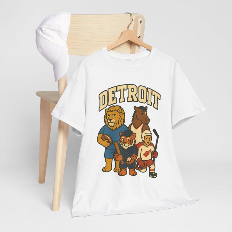 May include: White t-shirt with a graphic of four cartoon mascots representing Detroit sports teams. The word "DETROIT" is printed across the top in gold lettering. A white baseball cap hangs on a wooden chair.