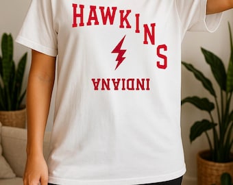 Puede incluir: Camiseta blanca con el texto rojo "HAWKINS INDIANA" y un rayo rojo. La camiseta de manga corta parece ser de algodón. La persona lleva pantalones cortos negros.