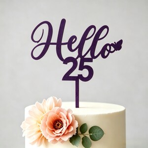 May include: A purple cake topper with the word "Hello" in cursive, the number "25", and a butterfly design. The topper is placed on a white cake decorated with peach-colored flowers and green leaves.