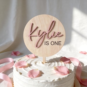 May include: A round wooden cake topper with the name "Kylie" in pink script and the words "IS ONE" in black block letters. The topper is placed on a white frosted cake decorated with pink rose petals.