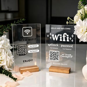 Könnte beinhalten: Zwei rechteckige Schilder aus klarem Acryl mit Holzsockeln. Das linke Schild zeigt ein WLAN-Symbol, den Text NEED WIFI?, einen QR-Code sowie Felder für Netzwerk und Passwort. Das rechte Schild zeigt HELLO AND WELCOME!, WiFi und einen QR-Code.
