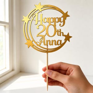 Può includere: Un cake topper dorato con le parole "Happy 20th Anna" ritagliate. Il design include una cornice circolare con stelle ed è attaccato a un bastoncino di legno.