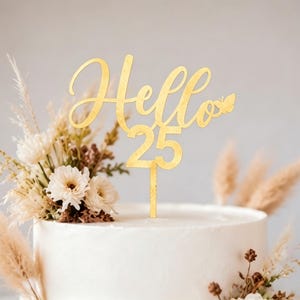 May include: A gold-colored cake topper with the words "Hello 25" in a script font, with a small butterfly detail. The topper is placed on a white frosted cake, decorated with dried flowers and wheat stalks.