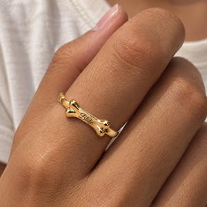 May include: A gold-toned ring featuring a central dog bone-shaped design. The surface of the bone is engraved with the name Blake in a cursive script. The band is thin and polished, encircling the finger with a smooth, reflective metallic finish.