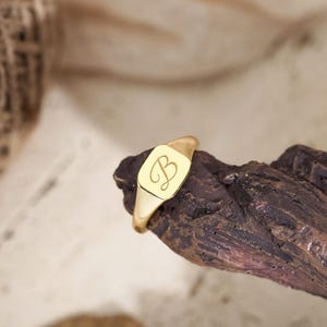 Peut inclure: Une bague chevalière dorée dotée d'une face rectangulaire plate gravée d'une lettre B en écriture cursive. La bague présente une finition métallique polie et un anneau lisse, mettant en valeur un design classique avec une initiale personnalisée.