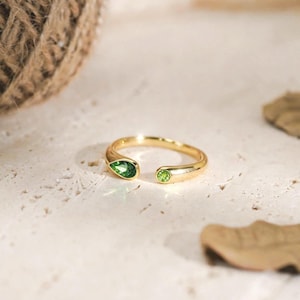 May include: A gold-toned open ring featuring two green stones. One stone is shaped like a teardrop, while the other is circular. The band is smooth and polished, with the stones set at each end of the opening, creating a distinct, balanced design.