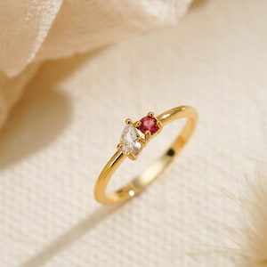 May include: A gold ring featuring two small stones set side by side. One stone is a clear, marquise-cut gem, and the other is a round, vibrant red gem. The band is thin and polished gold. The text JewelleryGiftCraft is visible at the bottom.