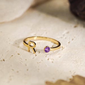 Op de afbeelding: Een goudkleurige open ring met een hoofdletter R aan het ene uiteinde en een kleine, ronde, paarse edelsteen aan het andere uiteinde. De ringband is dun en metaalachtig, wat het ontwerp met de letter en de levendige violette steen benadrukt.