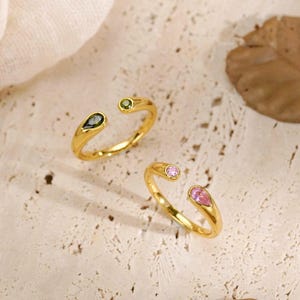 Puede incluir: Dos anillos abiertos de tono dorado. Un anillo presenta una pequeña piedra redonda verde y una piedra verde oscuro en forma de lágrima. El segundo anillo tiene una pequeña piedra redonda rosa y una piedra rosa en forma de lágrima. Acabado pulido.
