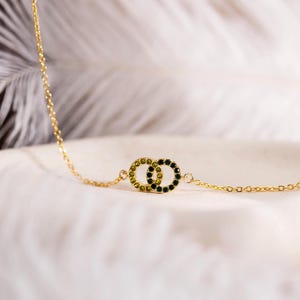 May include: A gold-toned chain necklace featuring two interlocking circular pendants. Each circle is embellished with small, dark green gemstones set along the entire circumference. The delicate metal chain connects to both sides of the linked rings.