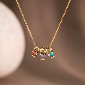 Puede incluir: Un collar de cadena dorada con tres pequeños dijes en forma de figuras estilizadas. Cada figura tiene un aro dorado en la parte superior y una piedra preciosa facetada en la base: una roja, una morada y una azul, colgando de la cadena.