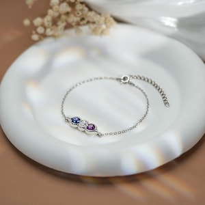 Op de afbeelding: Een delicate zilveren armband met drie edelstenen: een blauwe saffier, een heldere diamant en een paarse amethist. De armband ligt op een wit, geschulpte schaal, met een zachte, neutrale achtergrond.