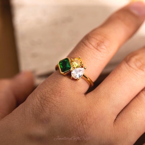May include: A gold-toned ring featuring three distinct gemstones: a rectangular emerald-green stone, a round yellow stone, and a pear-shaped clear white stone. The stones are set in a cluster arrangement on a thin, polished gold-coloured metal band.