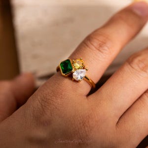 Puede incluir: Un anillo de oro con un grupo de piedras preciosas. El anillo tiene una piedra verde cuadrada con talla esmeralda, una piedra amarilla redonda y una piedra transparente en forma de pera. El anillo está en un dedo.