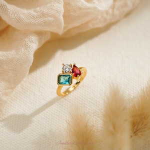 May include: A gold ring featuring a cluster of colorful gemstones. The ring has a rectangular teal stone, a round clear stone, and a pear-shaped red stone. The ring is set against a soft, neutral background.