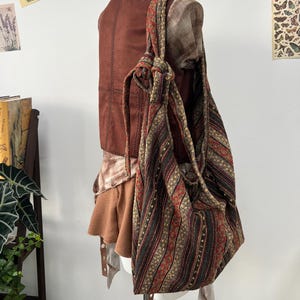 以下が含まれることがあります： 茶色と赤色の模様が入ったトートバッグがマネキンに掛けられています。バッグには結び目のついたハンドルと、茶色、赤、金色の縦縞模様があります。マネキンは茶色のベストと重ね着をしています。