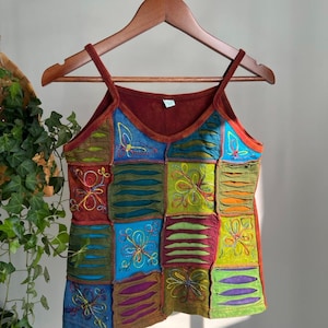 May include: A colourful patchwork camisole top with embroidered floral patterns. The top is made up of squares in shades of blue, green, orange and red. It hangs on a wooden hanger.