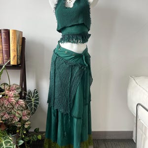 May include: A dark green two-piece outfit consisting of a sleeveless halter top with a frayed hem and a matching wrap skirt. The skirt features a textured fabric panel, a waistline with metallic buttons, and a contrasting light green lace trim hem.