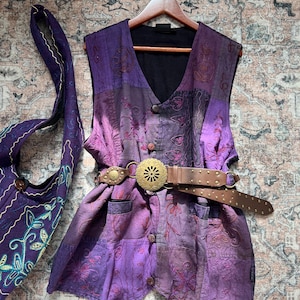 Puede incluir: Un chaleco morado sin mangas con paneles de patchwork, bordados intrincados y cierre de botones frontal. Un cinturón de cuero marrón con una hebilla grande, circular y ornamentada de metal dorado está sujeto a la cintura. Un bolso morado a juego está al lado.